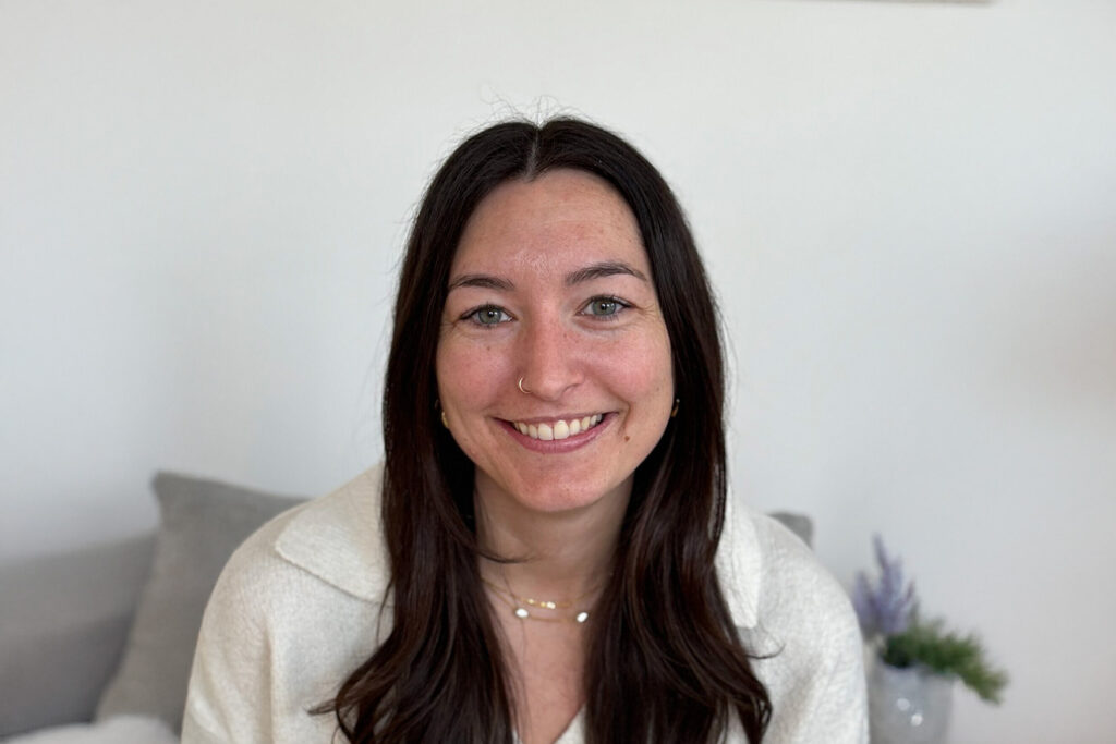 A smiling woman with light skin with a few freckles, long straight brown hair, and blue eyes. She is wearing a white shirt with a collar and has a nose ring on the left.