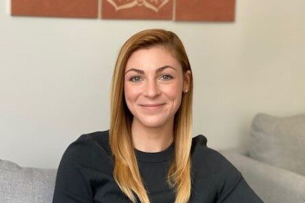 A smiling woman with light skin, straight blonde hair, and blue eyes. She is wearing a grey sweater.