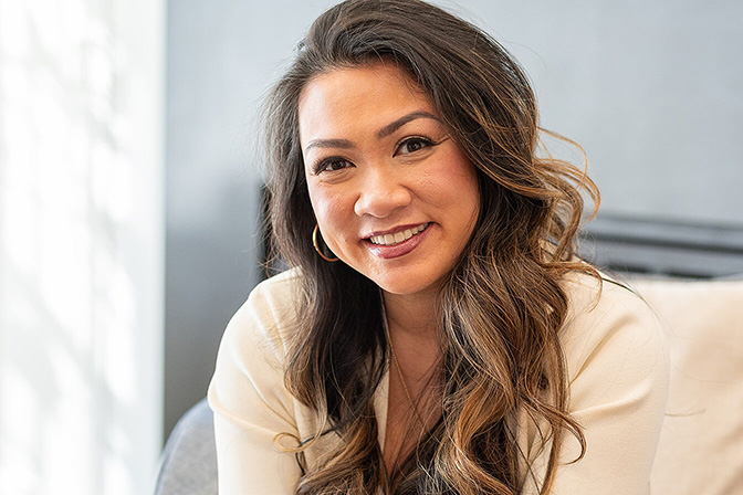 A smiling woman with medium skin, light brown wavy hair, and brown eyes. She is wearing a white blouse, hoop earrings, and burgundy lipstick.