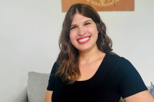 Smiling woman with light skin, wavy brown hair, and blue eyes. She is wearing a black top and red lipstick.