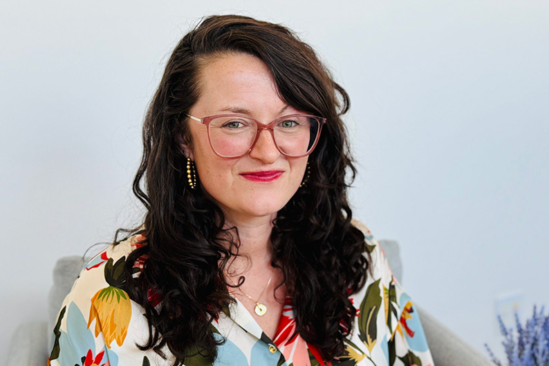 A smiling woman with light skin, long curly brown hair, and blue eyes. She is wearing a colorful flower-print blouse, and big glasses with a transparent red frame. She is also wearing hoop earrings and red lipstick.
