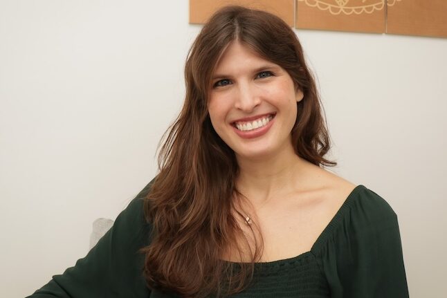 Smiling woman with light skin, wavy light brown hair, and blue eyes. She is wearing a forest green top and a thin gold necklace.