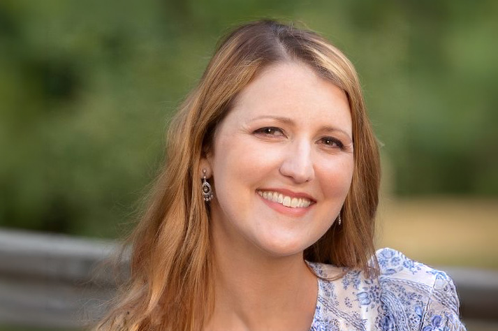 A smiling woman with light skin, losely waved and long blonde hair, and brown eyes. She is wearing a white blouse with a blue patterned design.